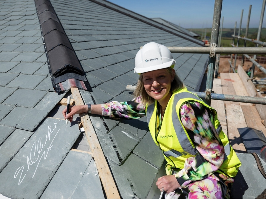 A woman with a Sanctuary branded hard-hat on and a high vis signing a roof tile on top of a building