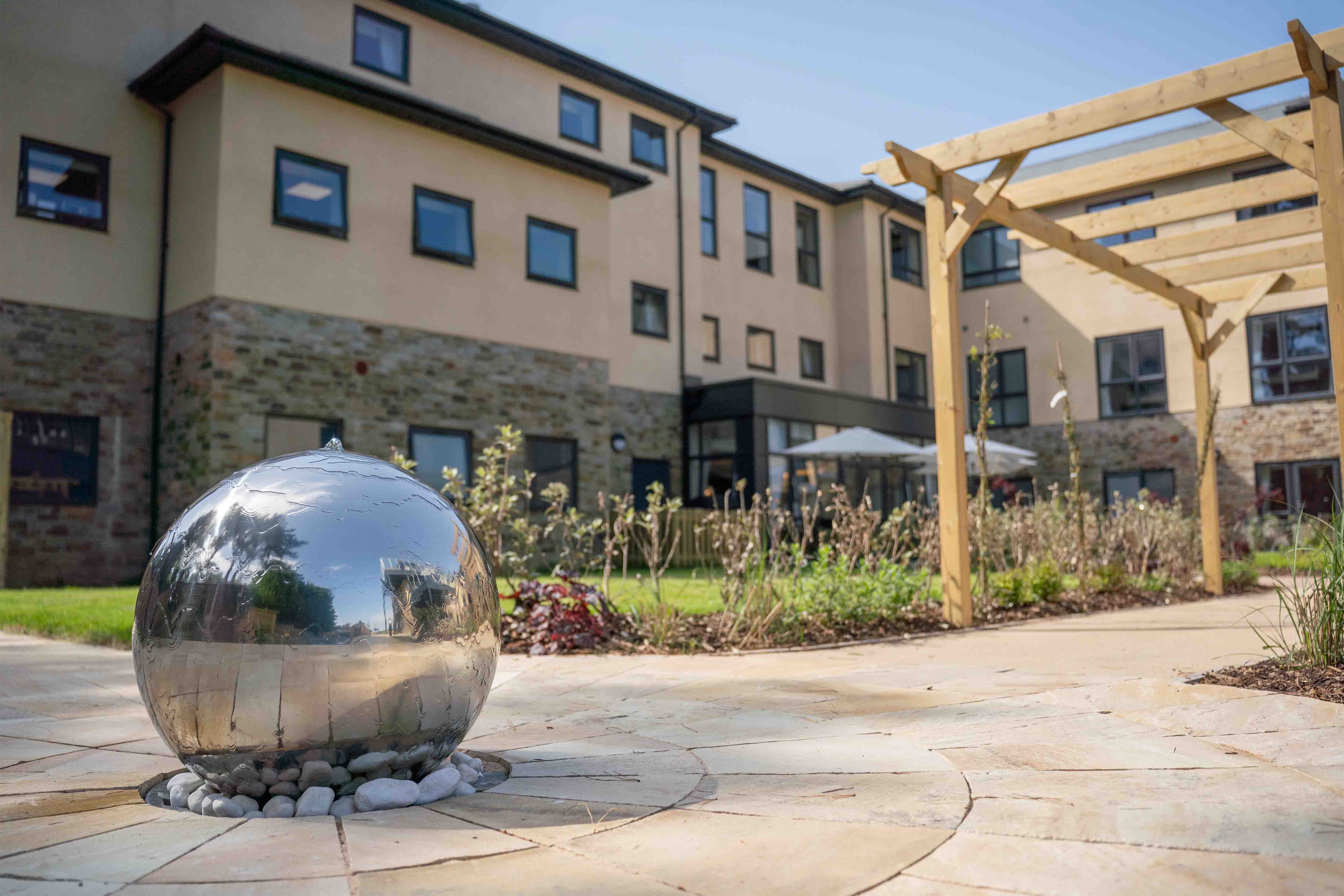 A stainless steel orb with a reflective surface sits on a paved area in front of a modern building. The building has a brick and stucco exterior with several windows and a wooden pergola in the background.
