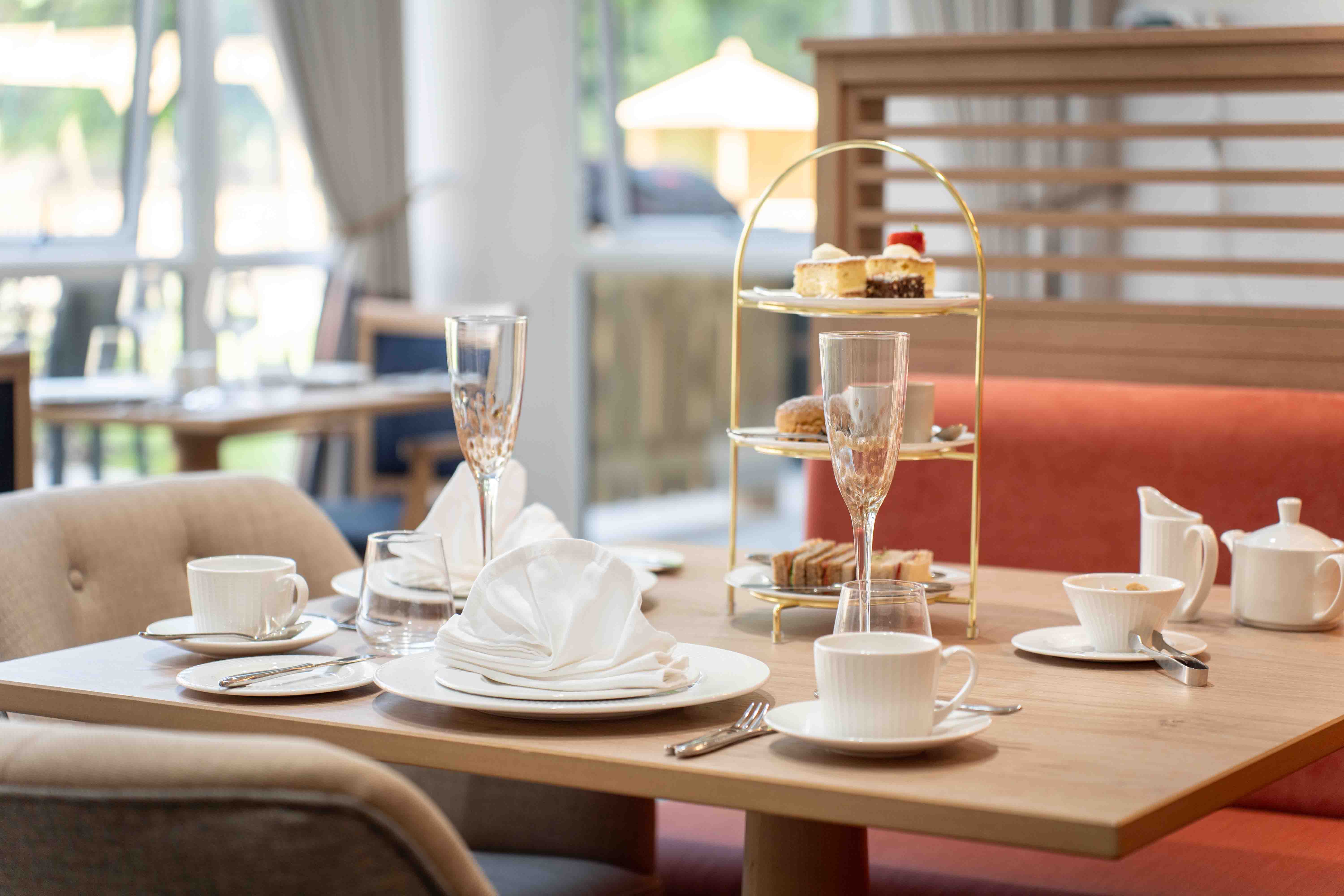 A table setting for afternoon tea, featuring a tiered stand with sandwiches and pastries, champagne, and delicate china.