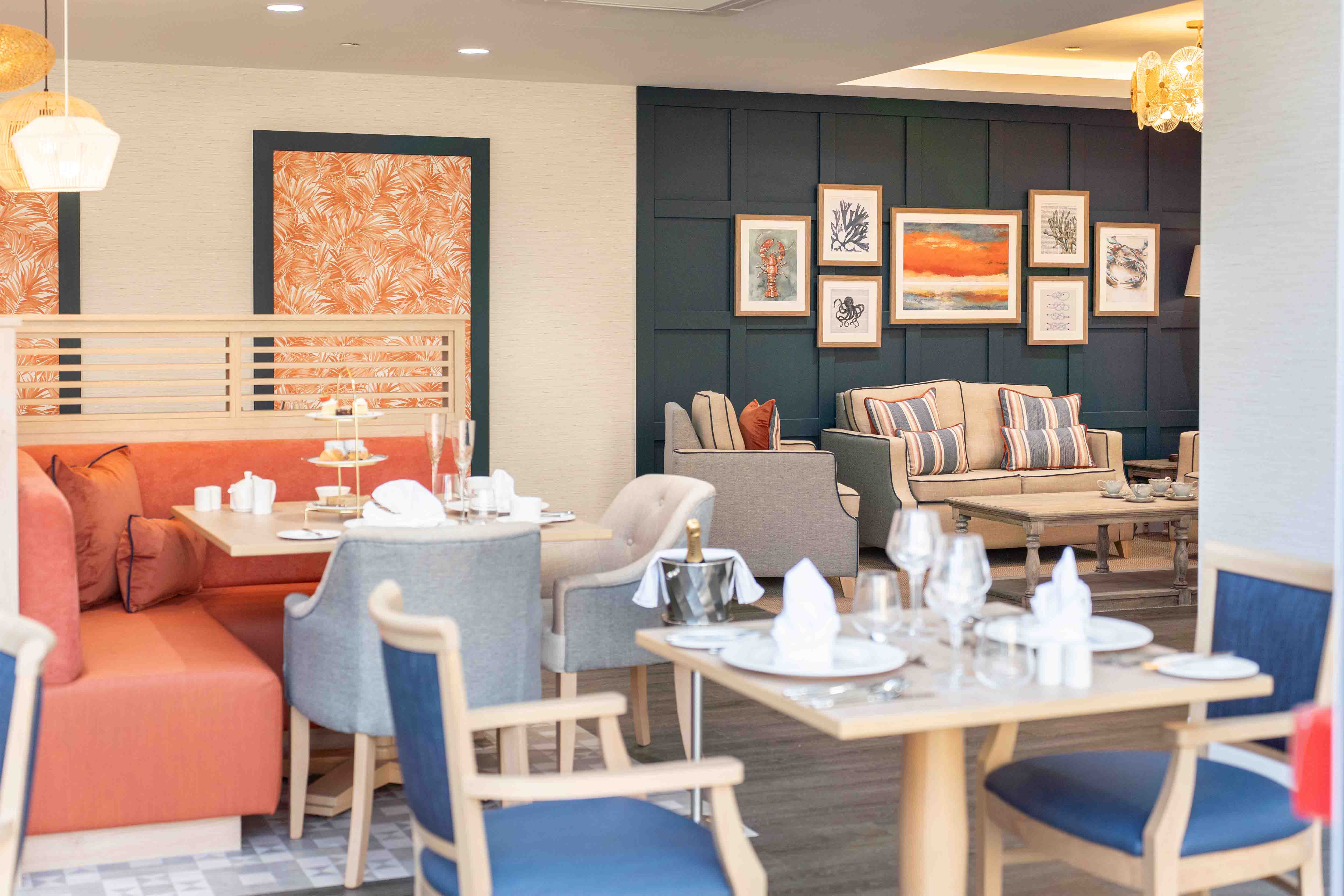 A warmly lit interior space features a set of tables with white tablecloths and glassware, arranged in a cozy seating area. A comfortable sofa and armchairs are positioned against a dark teal wall adorned with framed artwork. Decorative lighting and patterned curtains complete the scene.