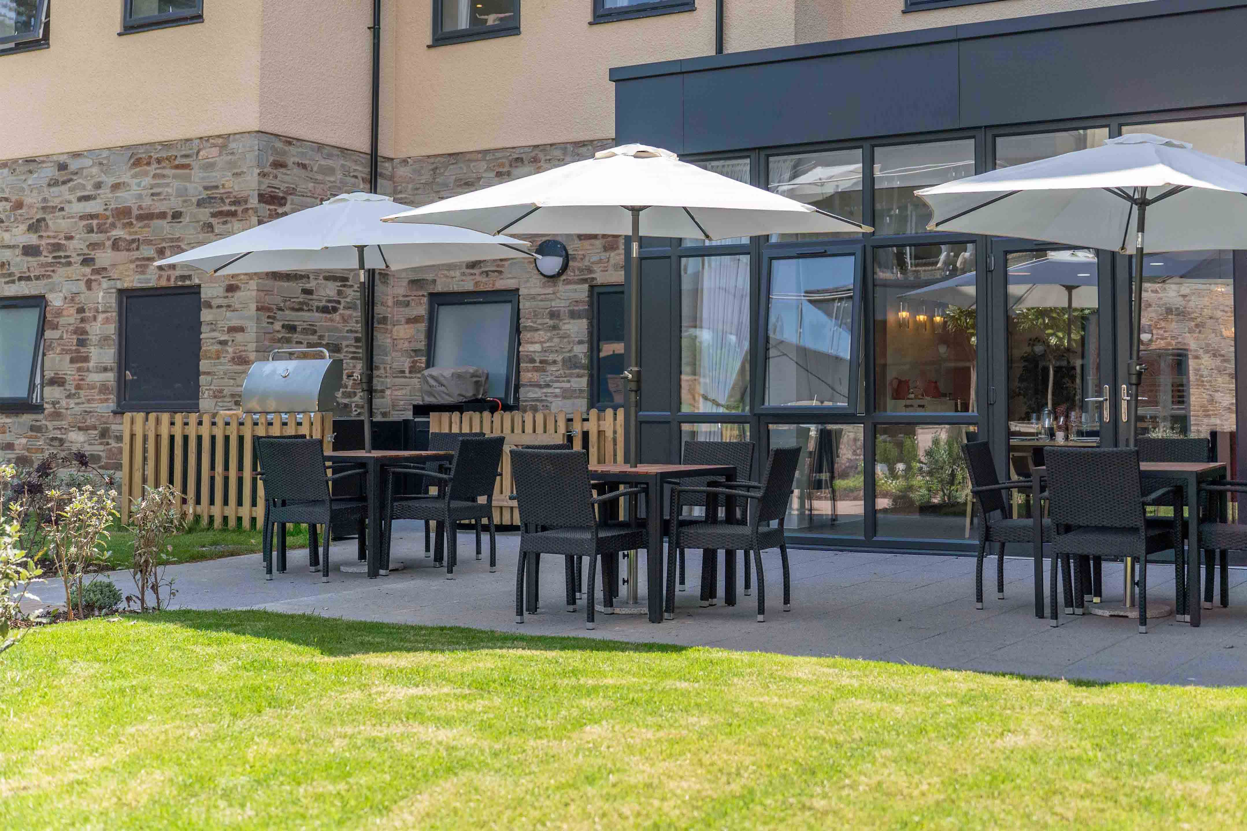 Outdoor patio with three white umbrellas and tables with chairs, adjacent to a building with large windows.