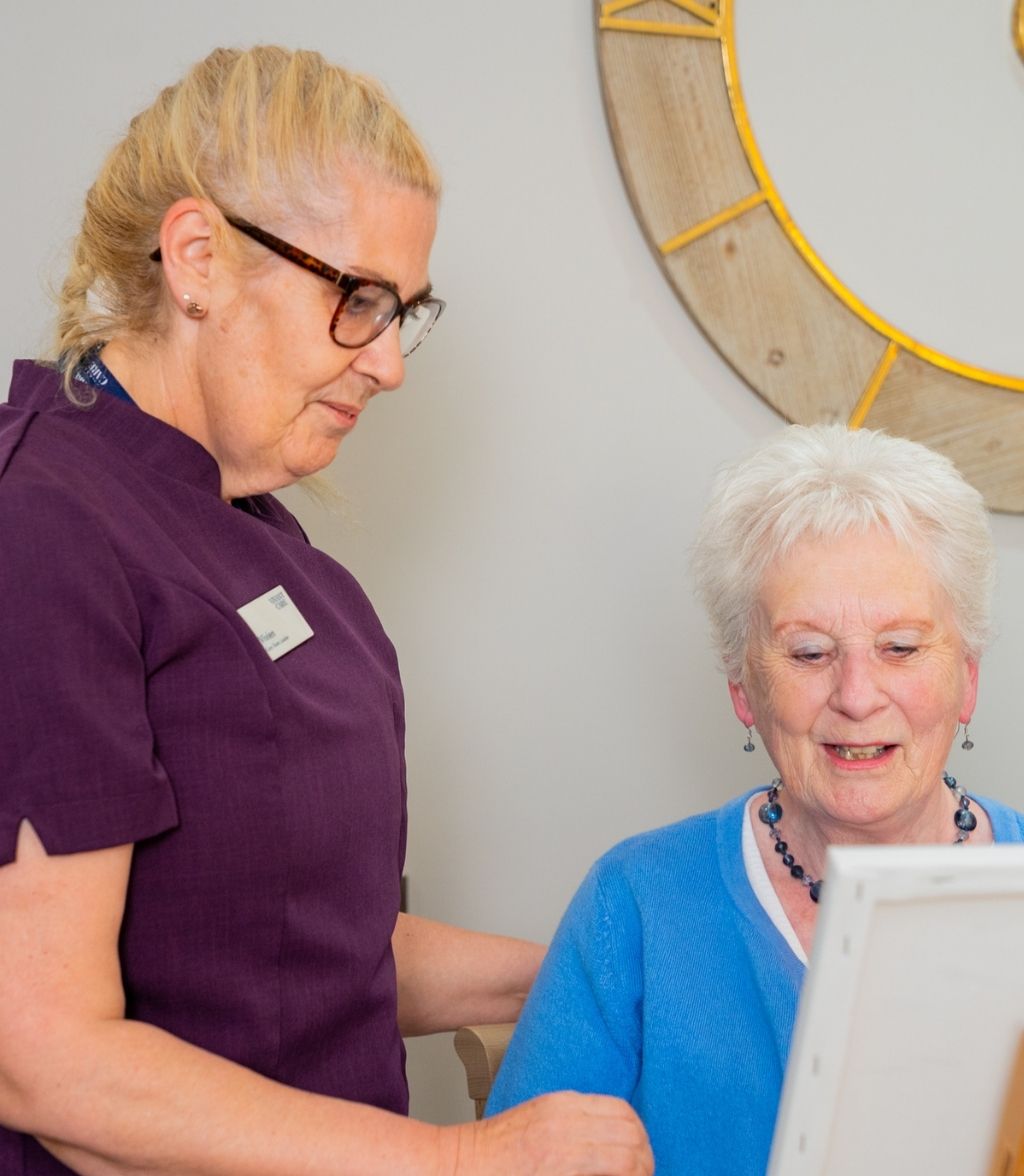 A woman in glasses and a purple shirt looks at an elderly woman wearing a blue sweater and a silver necklace.
