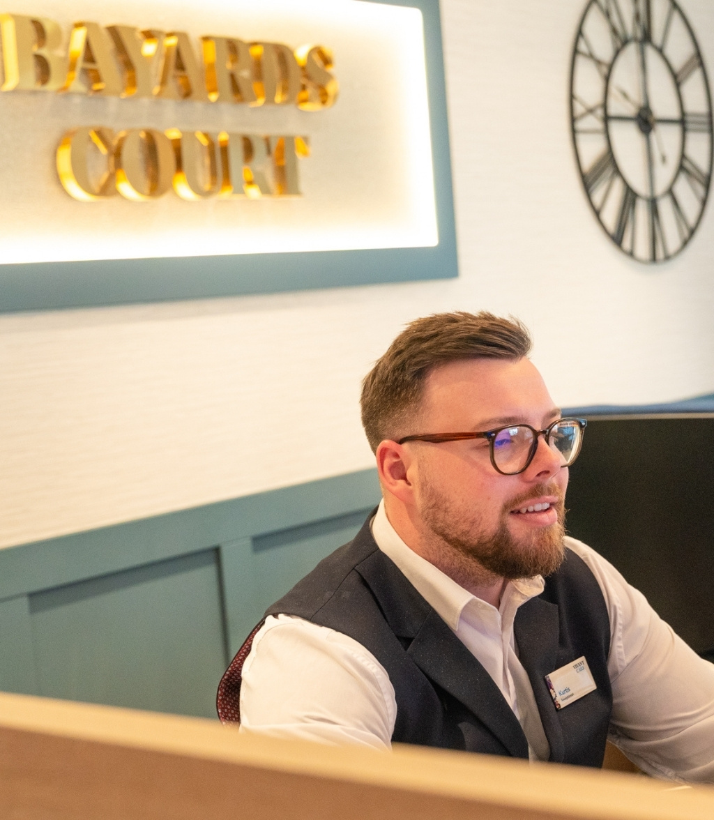 “Bayards Court” signage is visible in the background. A man wearing glasses and a waistcoat is seated at a reception desk, smiling slightly at the camera. A large, ornate clock is mounted on the wall behind him.