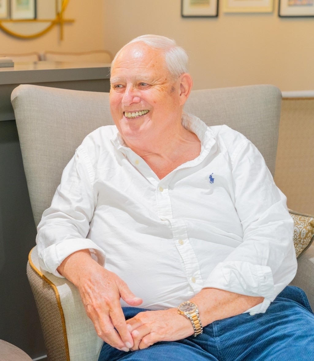 A smiling elderly man with white hair and a gold wristwatch is seated in a gray upholstered armchair. He is wearing a white polo shirt and blue jeans. A decorative gold object is visible in the background, along with framed artwork on the wall.