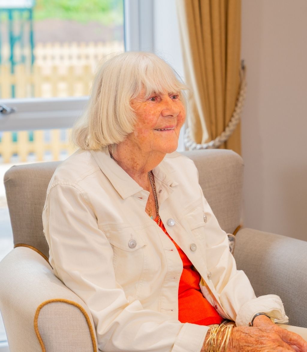 A smiling elderly woman with short, blonde hair is seated in a grey armchair. She is wearing a white denim jacket over an orange shirt and a pair of gold bracelets. A window with a garden view is visible in the background.