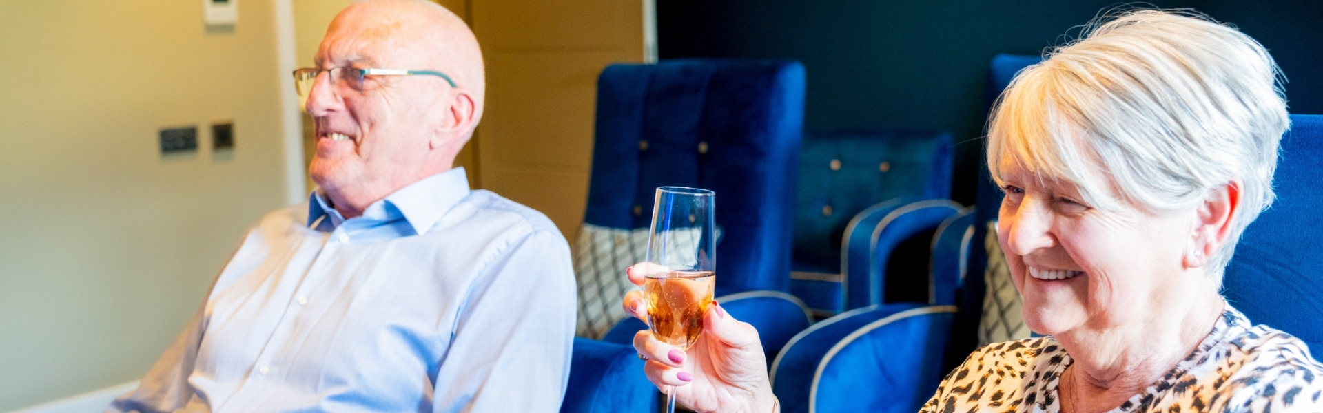 An elderly man and woman are seated in blue armchairs, smiling and raising champagne glasses in a toast. Behind them are three framed artwork pieces depicting stylized floral designs.