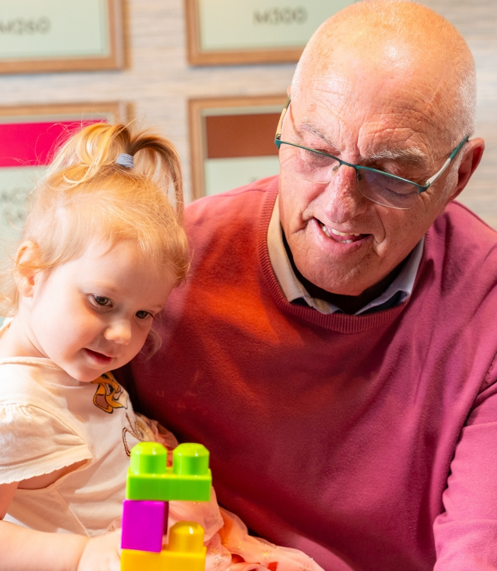 A smiling elderly man with glasses is intently looking at a young girl with blonde hair in a ponytail as she plays with a set of colorful building blocks.