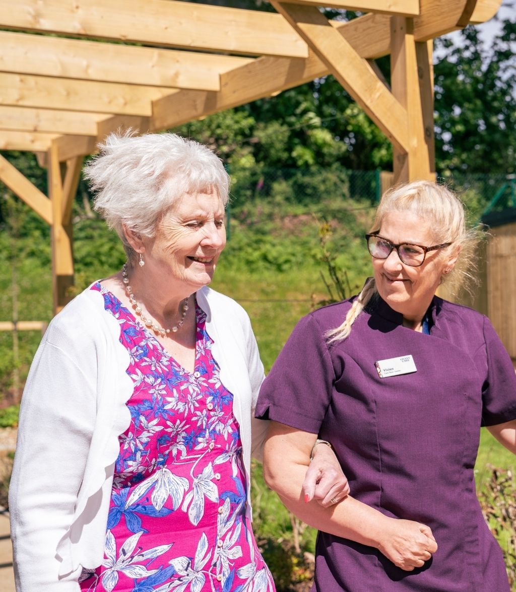 A smiling elderly woman with short white hair and a pink and blue floral shirt walks alongside a healthcare professional wearing a purple tunic and glasses. They are standing under a wooden pergola in a garden setting.