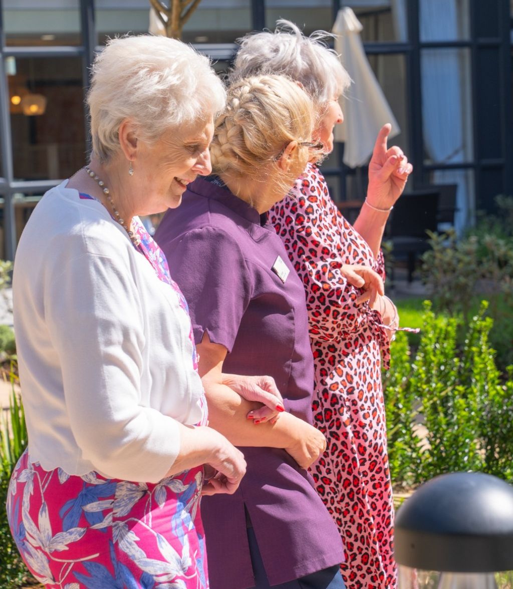 Two elderly women are walking outdoors, one in a pink and white floral shirt and the other in a red and white leopard print dress. They are both looking up and gesturing with their hands.